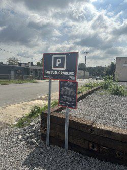 A "Paid Public Parking" sign stands on gravel by a road under a partly cloudy sky, with buildings and power lines visible in the background.