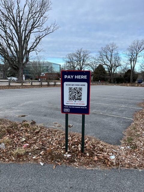 A "Pay Here" sign with a QR code stands in front of an empty parking lot with bare trees and scattered leaves on the ground. Buildings are visible in the background under a cloudy sky.