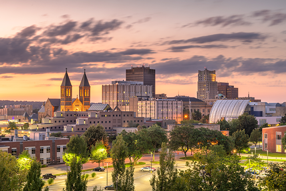 Cityscape at sunset showing a mix of historic and modern buildings, including twin-spired church towers, office buildings, and trees in the foreground under a partly cloudy sky.