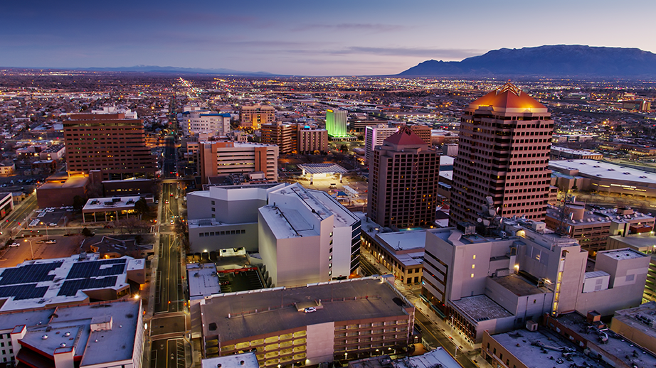 Aerial view of downtown Albuquerque, New Mexico at dusk, with tall buildings, city lights, and mountains visible in the distance under a purple sky.