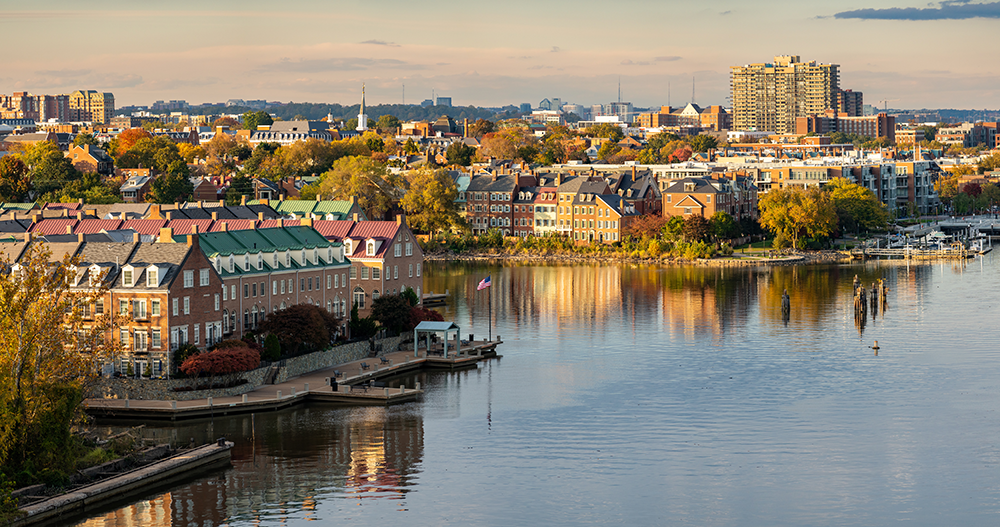 A scenic waterfront view of Old Town Alexandria, Virginia, with colorful historic townhouses, trees with autumn foliage, and calm water reflecting the buildings at sunset.