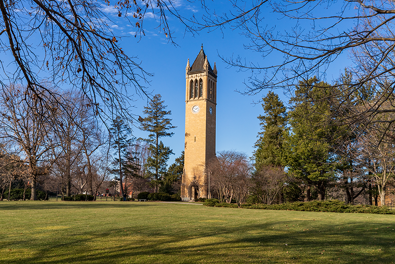 A tall brick clock tower stands on a grassy lawn surrounded by trees with bare branches under a clear blue sky.