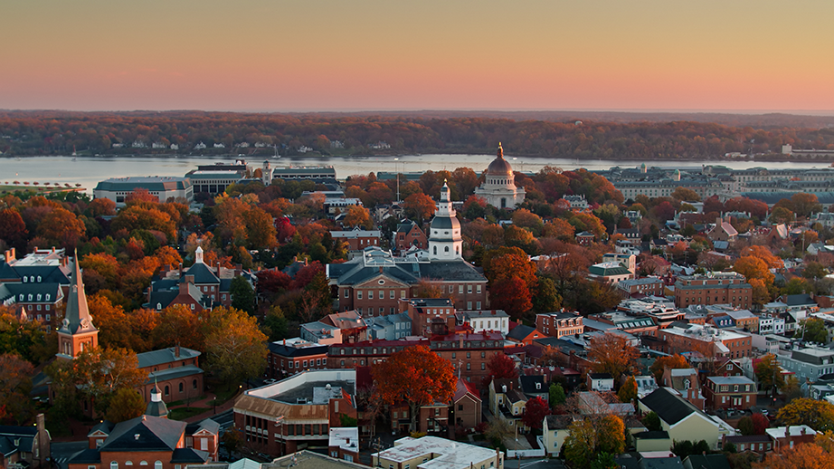 Aerial view of a historic town with colorful autumn trees, brick buildings, and a prominent domed capitol building at sunset, with a river in the background.
