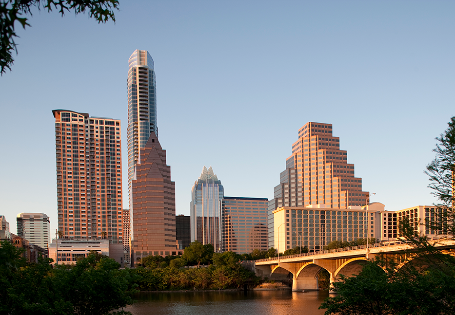 Downtown city skyline with tall modern and stepped buildings, a bridge over a river in the foreground, and trees along the water, all lit by warm sunlight.