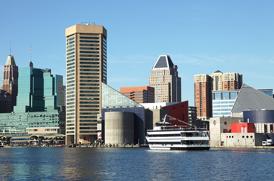 Baltimore Inner Harbor skyline with modern and historic buildings, a docked white and blue boat, and clear blue sky reflecting on the water.