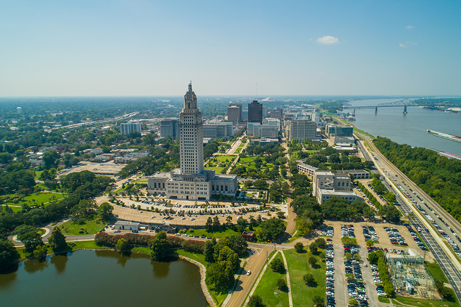 Aerial view of downtown Baton Rouge, Louisiana, featuring the tall Louisiana State Capitol building, surrounding green areas, and the Mississippi River to the right.