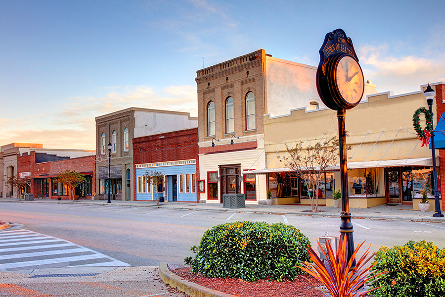 A quiet small-town main street at sunset, lined with historic brick buildings and shops. A vintage clock stands on the sidewalk near tidy landscaping and a crosswalk. The street is empty and calm.