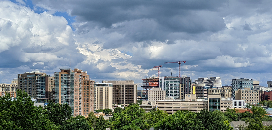 A cityscape featuring modern buildings and construction cranes under a dramatic, cloudy sky, with green trees in the foreground.