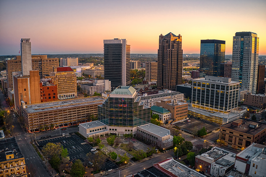 Aerial view of downtown Birmingham, Alabama at sunset, featuring tall office buildings, a glass-roofed structure, parking lots, and city streets with a clear sky in the background.