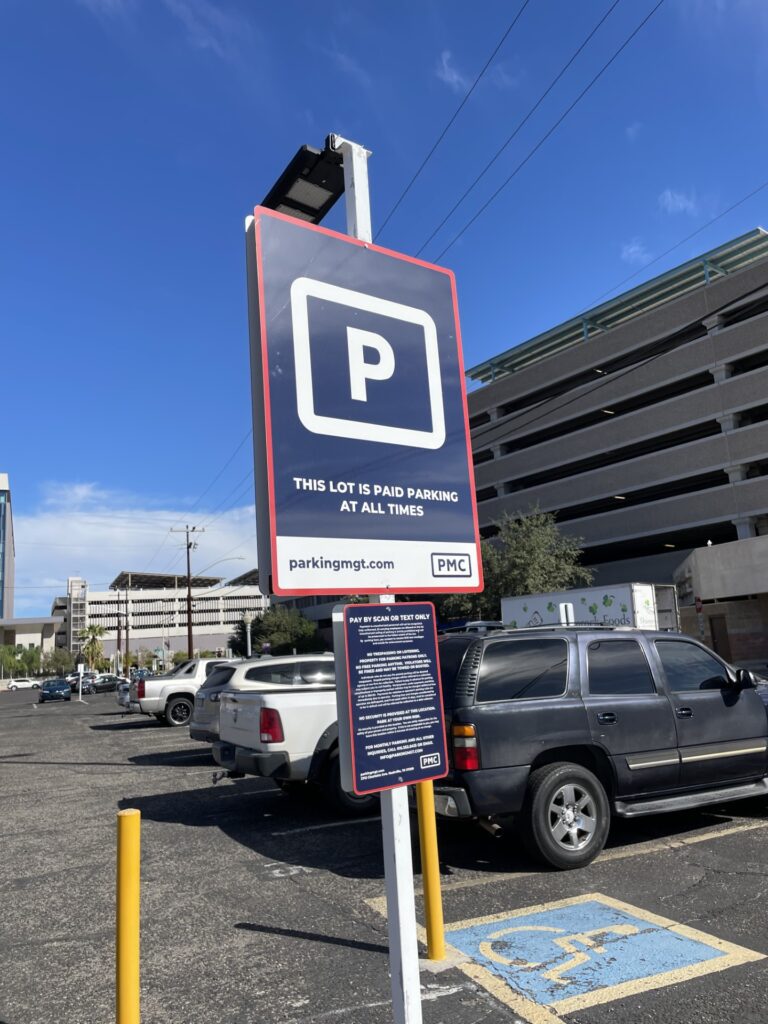 A large PMC parking sign in a lot reads "This lot is paid parking at all times" with a white "P" on a dark blue background and red border. Cars are parked nearby and a multi-level parking garage is visible in the background.