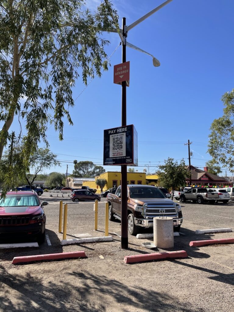 A parking lot with several cars and a PMC sign on a pole that reads "PAY HERE" with a QR code for payment. The sky is clear and blue, with trees and buildings in the background.
