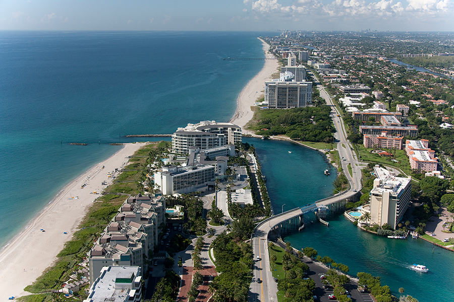 Aerial view of a coastal city with high-rise buildings and resorts along a sandy beach, a bridge crossing a blue waterway, and the ocean extending to the horizon under a partly cloudy sky.