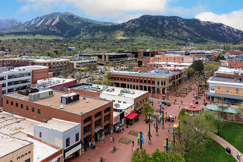 Aerial view of a vibrant downtown area with brick buildings, pedestrian walkways, people walking, green trees, and mountains in the background under a partly cloudy sky.