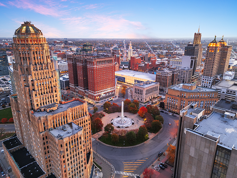 Aerial view of downtown Buffalo, New York at sunrise, featuring historic buildings, a roundabout with a fountain, and clear skies above the city.