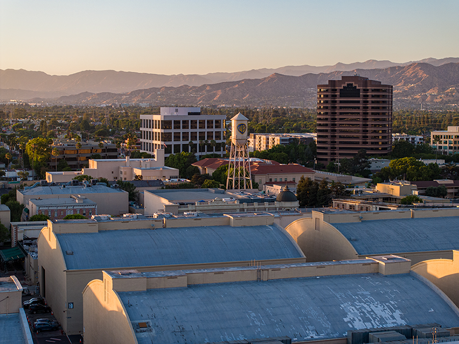 A cityscape at sunset featuring a white water tower with a studio logo, office buildings, and mountains in the background. The lighting casts a warm glow over the urban scene.