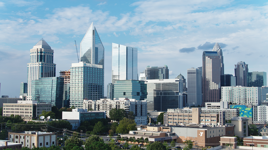 A cityscape showing modern skyscrapers and office buildings under a partly cloudy sky, with smaller buildings and trees in the foreground.