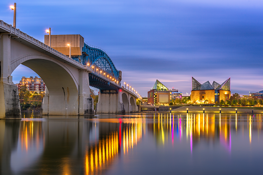 A bridge with lights spans a calm river at dusk, reflecting colorful lights from buildings, including a glass pyramid and angular structures, along the shoreline.