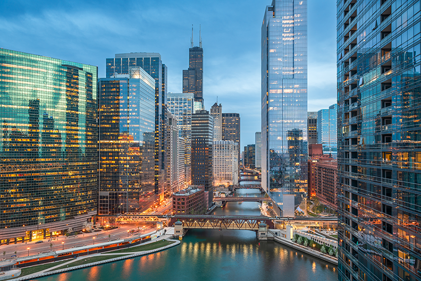 View of downtown Chicago at dusk, with tall modern skyscrapers reflecting lights, the Chicago River running through the center, and bridges crossing over the water. The scene is bright and vibrant with city life.