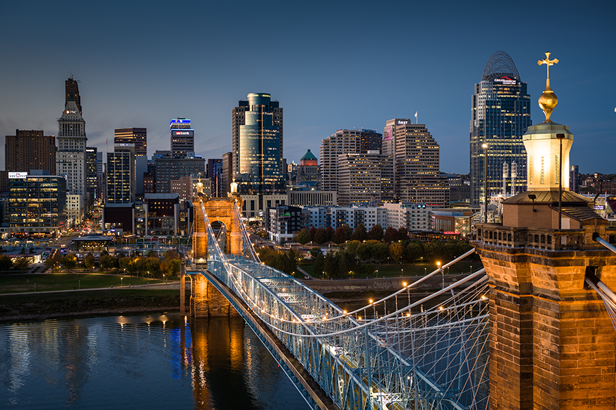 A city skyline at dusk features tall buildings with lights on, a blue suspension bridge crossing a river, and a golden sculpture on a nearby stone structure in the foreground.