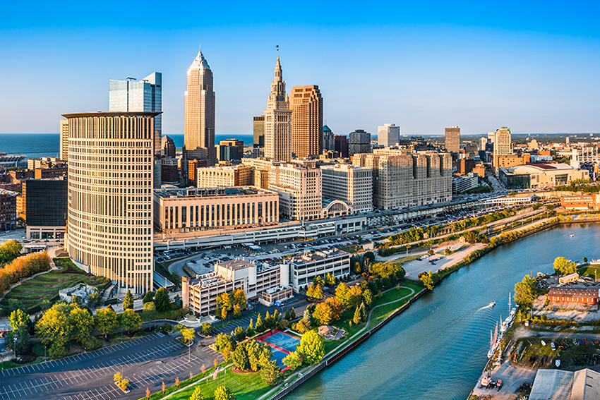 Aerial view of downtown Cleveland, Ohio, showing modern and historic skyscrapers, a river bordered by trees, a curved road, and a sunny, clear blue sky.