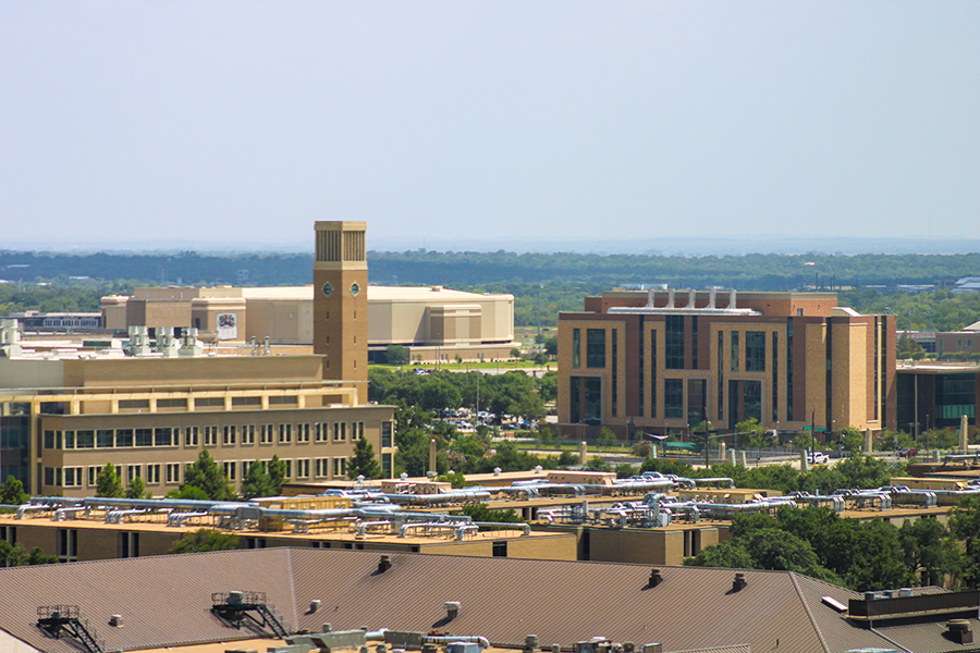 An elevated view of a university campus with several modern buildings, including a tall clock tower and tree-lined streets. Buses are parked along the roads, and the landscape stretches to the distant horizon.