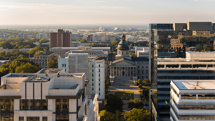 A cityscape view of downtown Columbia, South Carolina, featuring the South Carolina State House with its dome, surrounded by modern office buildings and trees under a clear sky.
