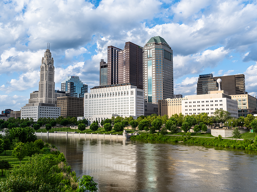 A city skyline with tall modern and historic buildings rises above a tree-lined riverbank on a partly cloudy day, with the buildings reflected in the calm water below.