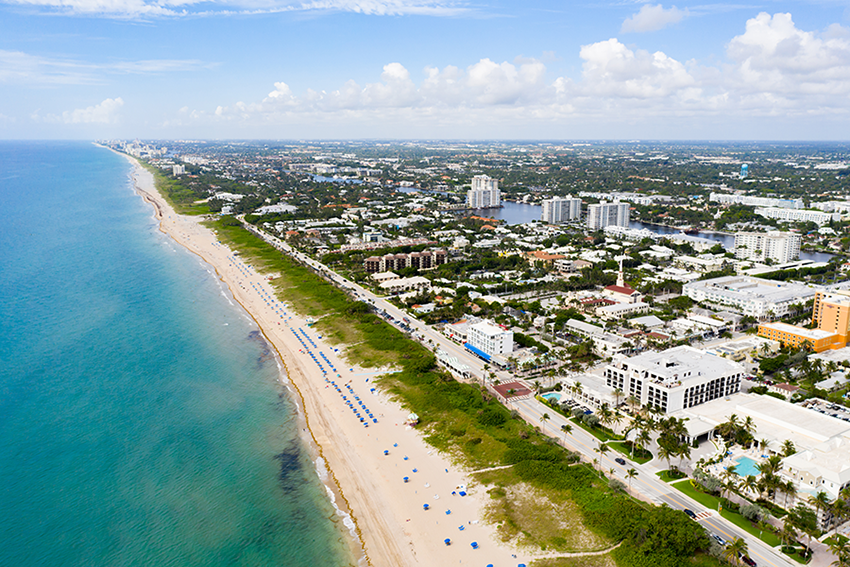 Aerial view of a coastal city with sandy beach, blue ocean, beach umbrellas, and numerous buildings and roads bordered by palm trees under a partly cloudy sky.
