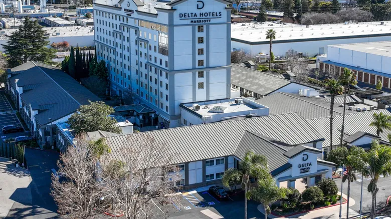 Aerial view of Delta Hotels by Marriott, featuring a multi-story white building with the hotel’s logo, surrounded by trees, smaller buildings, and nearby industrial structures.