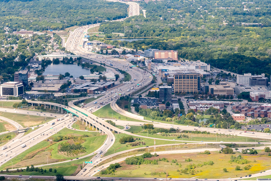 Aerial view of a city with multiple highways intersecting, surrounded by buildings, trees, and residential areas, with dense green forest in the background under a clear sky.