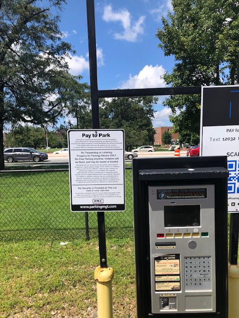A parking payment machine stands in front of a fenced grassy area. A sign above reads “Pay to Park” with instructions. Cars are visible on the road behind the fence, and trees and a building are in the background under a blue sky.