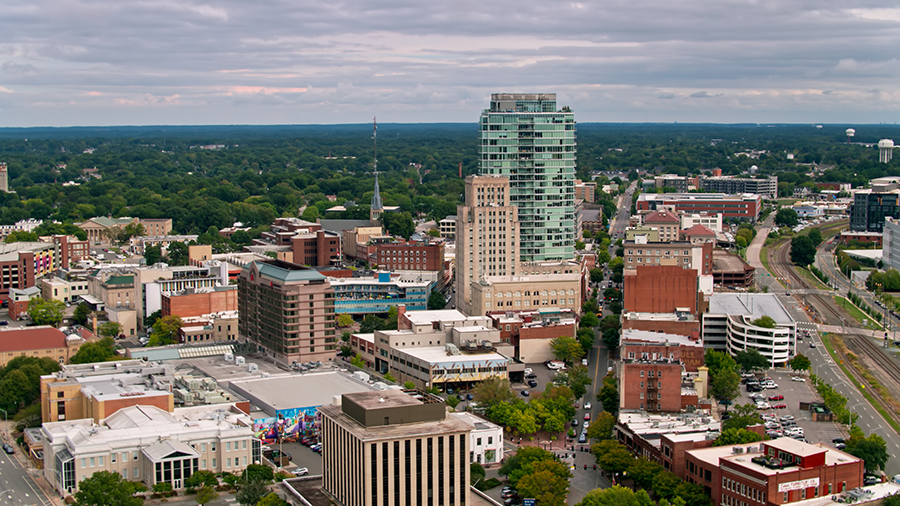 Aerial view of downtown Raleigh, North Carolina, showing a mix of mid-rise buildings, green trees, and city streets under a cloudy sky.