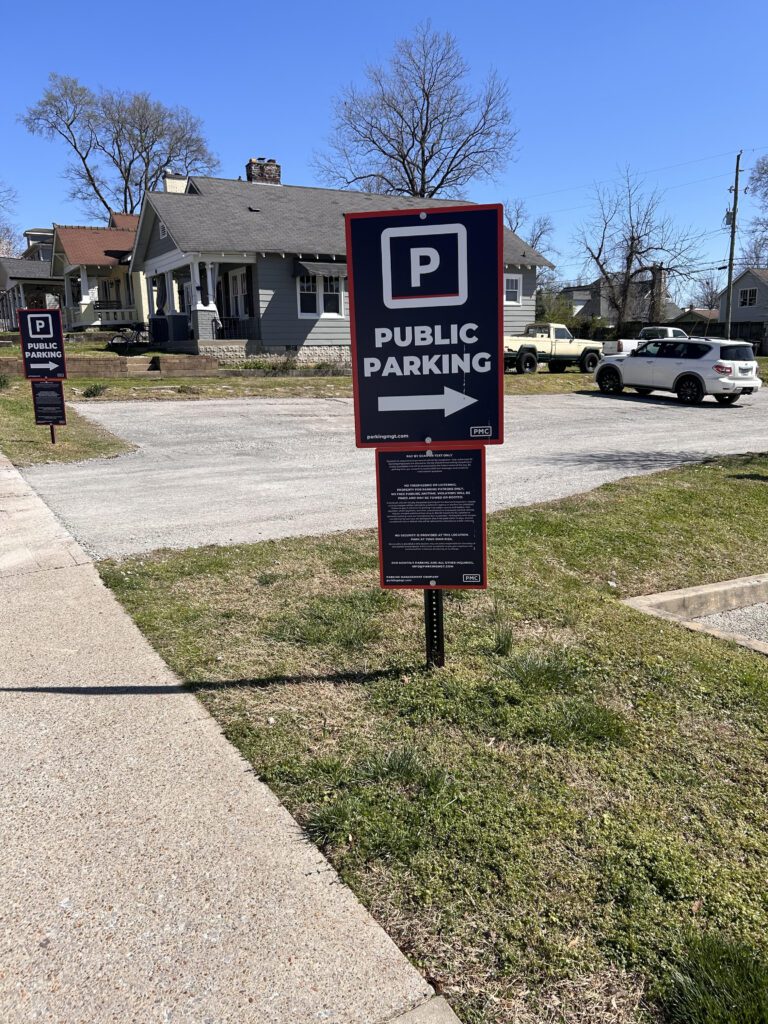 A sign on grass near a sidewalk reads "Public Parking" with a large "P" symbol and an arrow pointing right. Houses, parked cars, and trees are visible in the background under a clear blue sky.