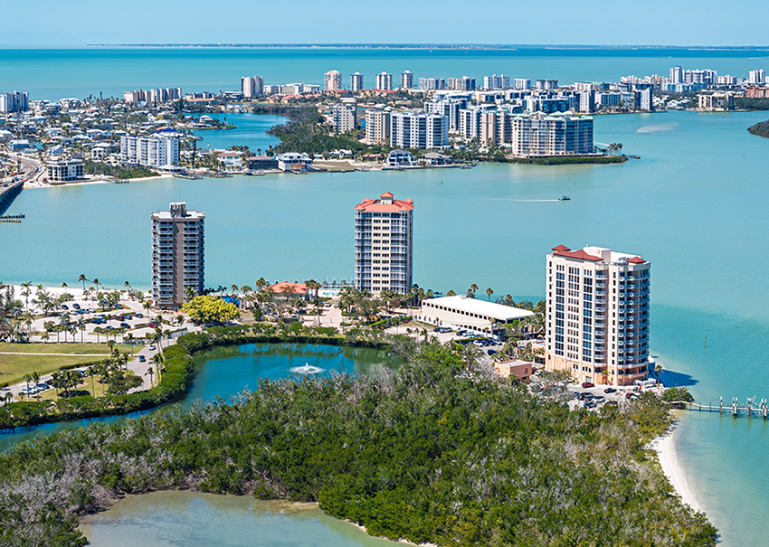 Aerial view of coastal city with tall modern buildings, blue-green water, small islands, and lush greenery in the foreground under a clear sky.