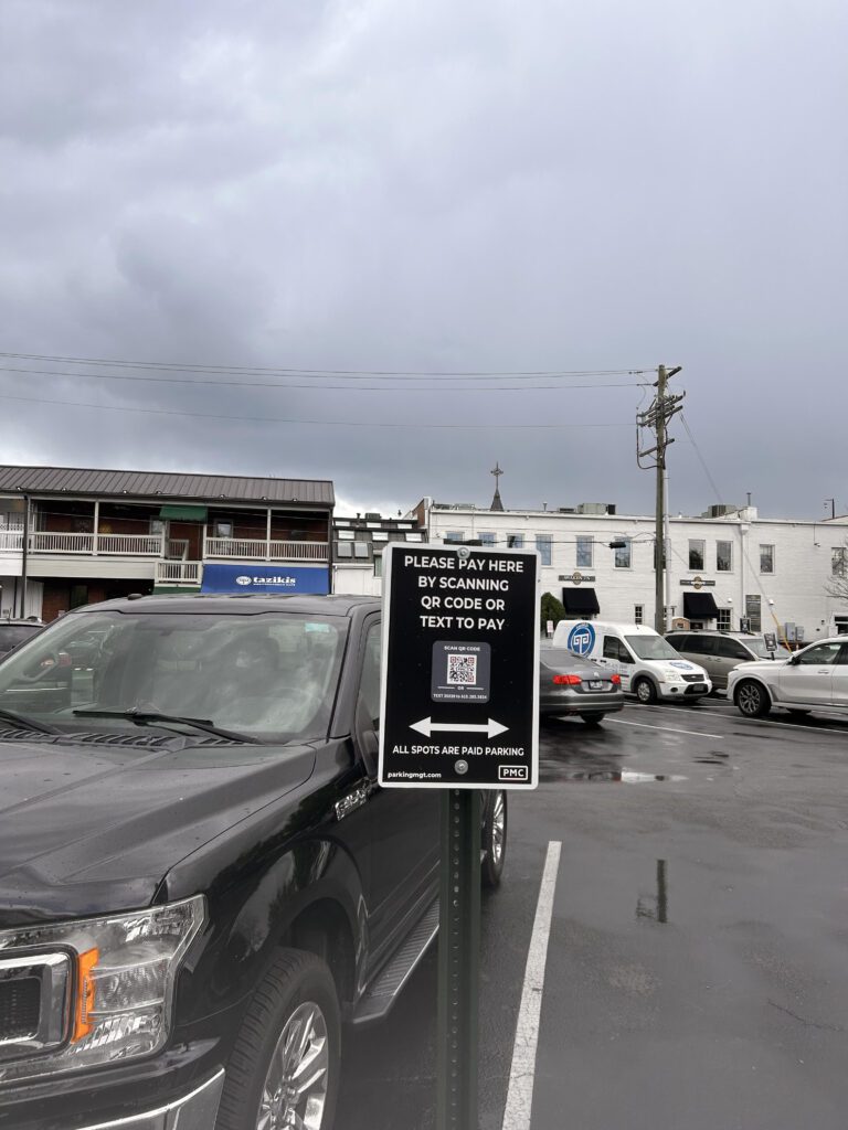 A parking lot on a cloudy day with a sign instructing people to pay by scanning a QR code or texting. Several parked cars and buildings are visible in the background.