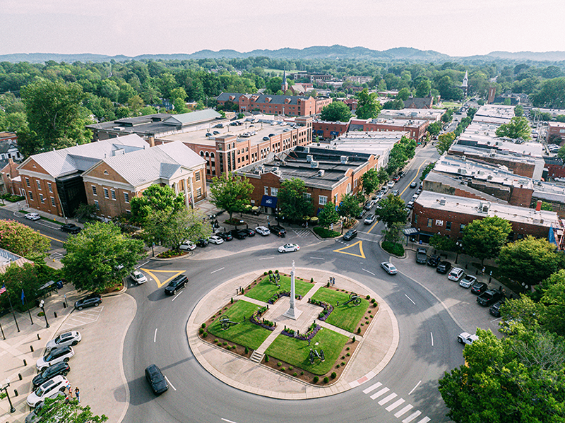 Aerial view of a small town square with a central roundabout featuring a monument, surrounded by buildings, trees, parked cars, and distant green hills under a hazy sky.