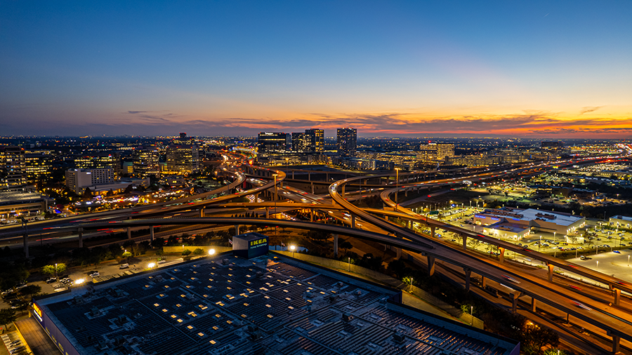 Aerial view of a sprawling highway interchange lit by streetlights at sunset, with city buildings and a shopping center in the background, and a colorful sky above the urban landscape.