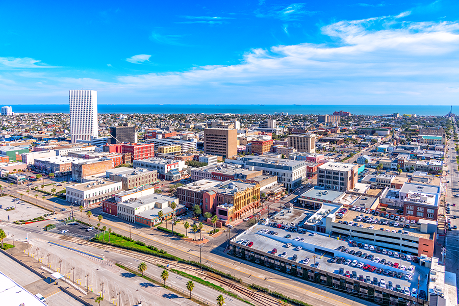 Aerial view of a coastal city with mid-rise buildings, parking lots, palm trees, and roads, extending toward a blue ocean under a bright, partly cloudy sky.