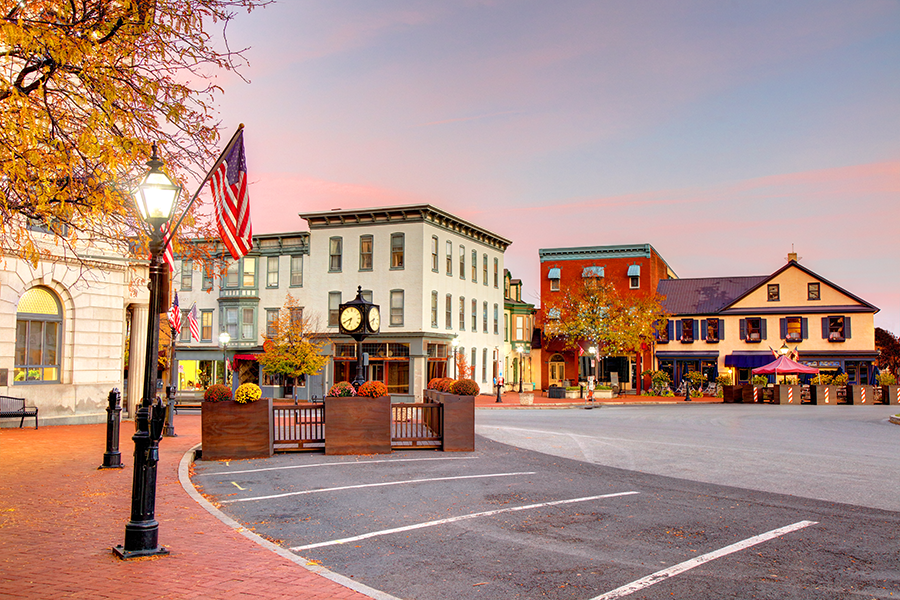A small town square at sunset with historic buildings, autumn trees, street lamps, and American flags. Outdoor seating and flower pots line the sidewalk, and the sky is tinted pink and orange.