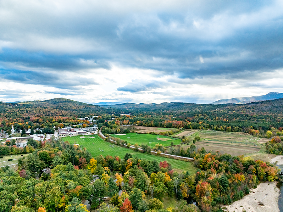 Aerial view of a rural landscape with green fields, scattered trees in autumn colors, a small town, and distant hills under a partly cloudy sky.