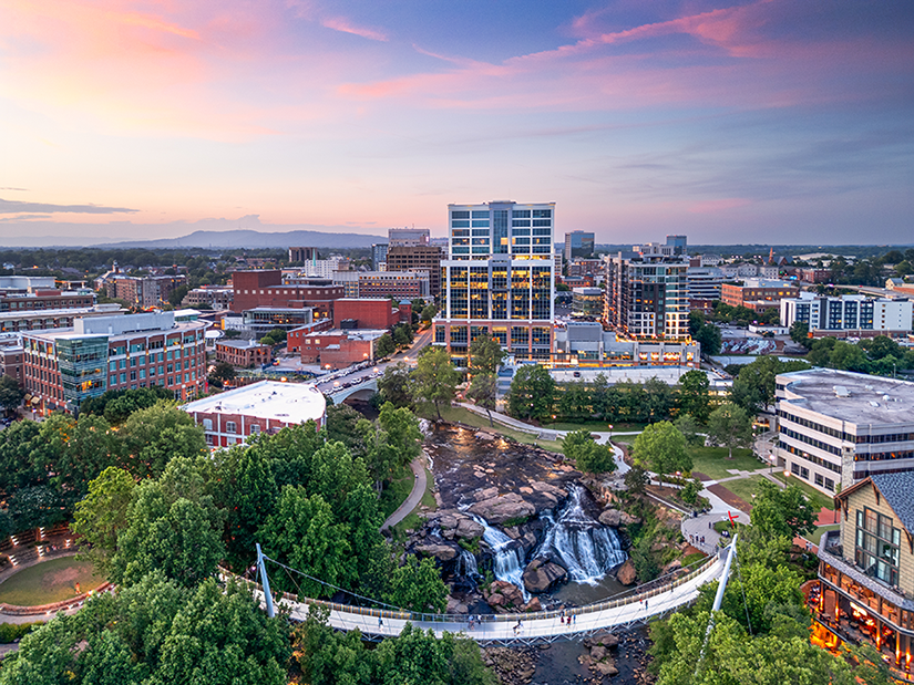 Aerial view of a city at sunset featuring a modern skyline, a park with a waterfall, and a curved pedestrian suspension bridge in the foreground. Trees and buildings are visible under a colorful sky.