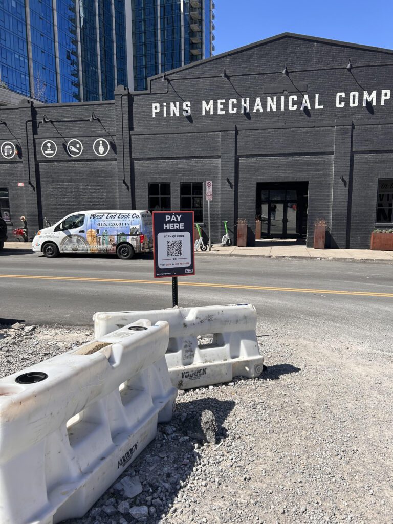 A "Pay Here" parking sign stands in front of white barriers on a gravel lot, with Pins Mechanical Company’s black building and a parked van visible across the street.