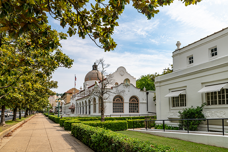 A wide sidewalk lined with green trees and neatly trimmed bushes runs alongside historic white buildings with arched windows and a domed roof under a partly cloudy sky.