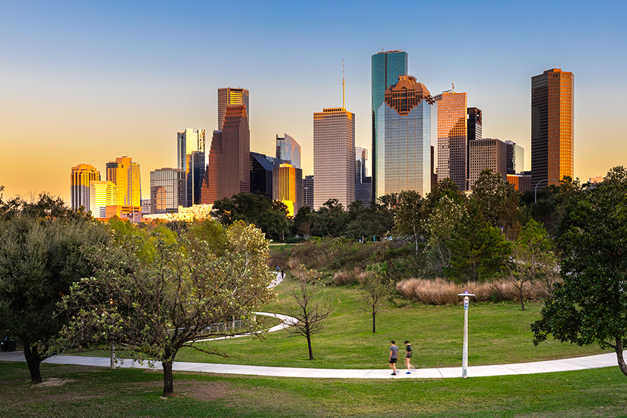 Two people walk on a path through a green park with trees, while the sun sets, casting a warm glow on the tall skyscrapers of a city skyline in the background.