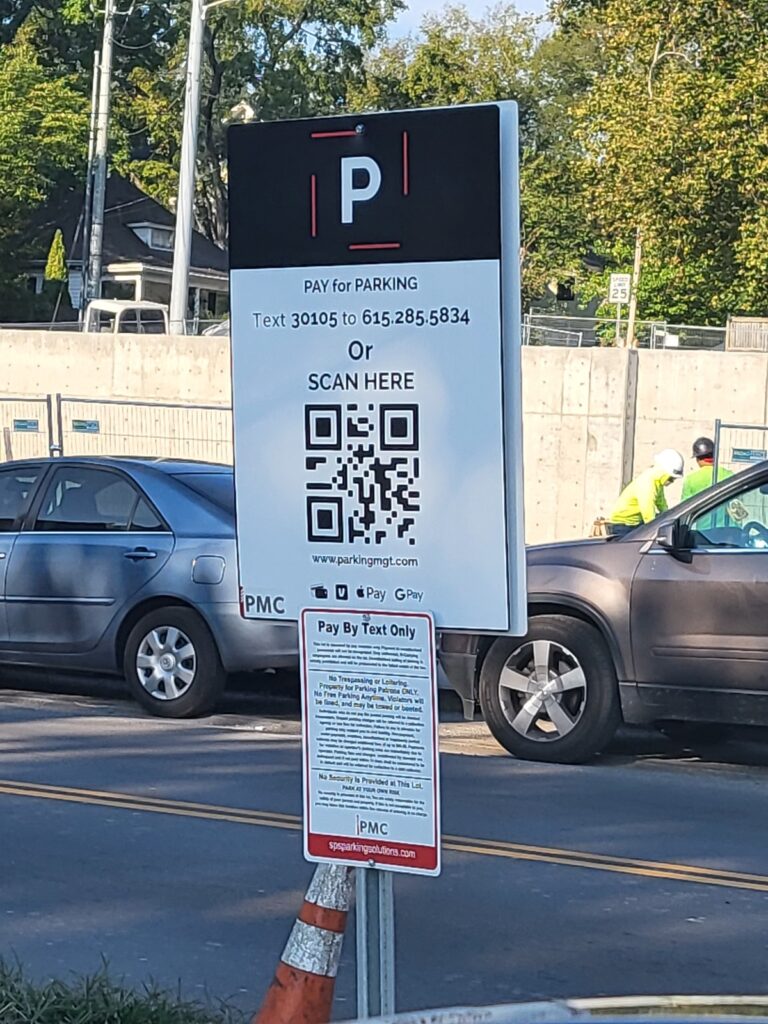 A parking sign instructs users to pay by texting a number or scanning a QR code. A car is parked nearby, and two people in safety vests are visible in the background near a road.