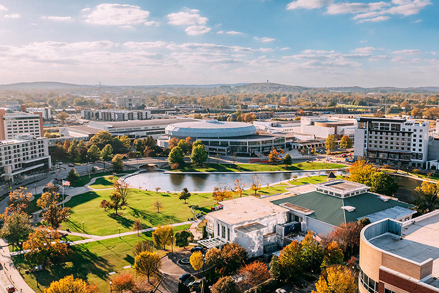 Aerial view of a modern university campus with green lawns, a small lake, several academic buildings, and tree-lined walkways, set against a backdrop of distant hills under a partly cloudy sky.