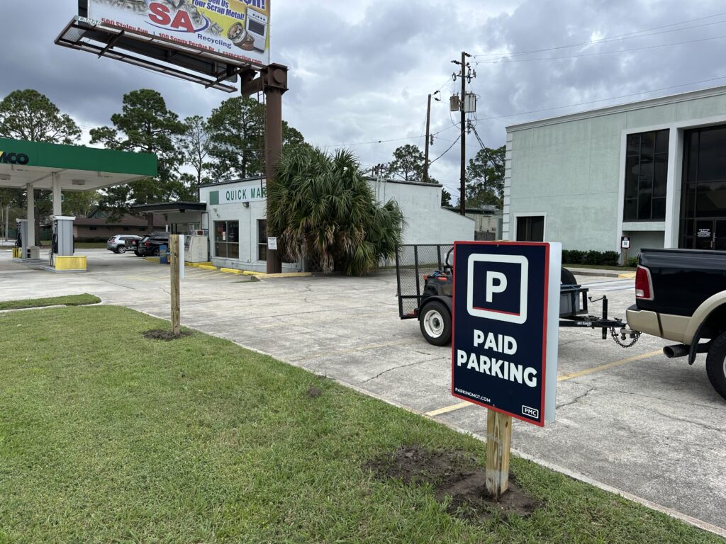 A PMC sign reading "Paid Parking" stands in a parking lot near a gas station and a Quick Mart store, with a pickup truck and trailer parked nearby.