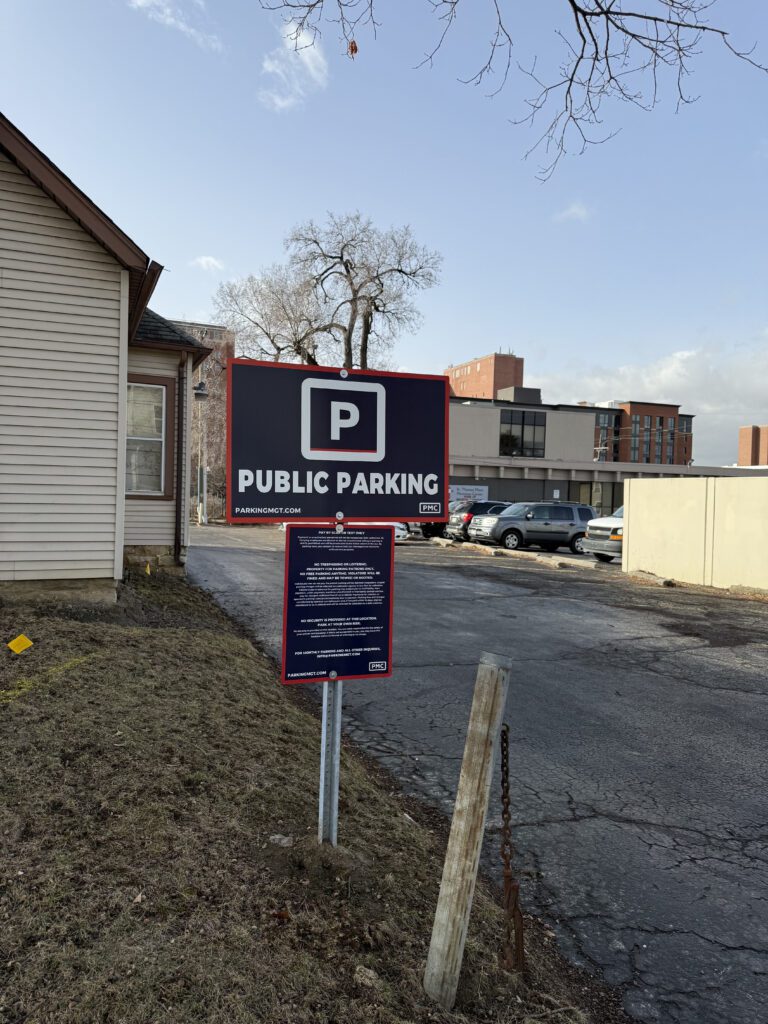 A sign reading "Public Parking" stands at the entrance to a small parking lot next to a beige building, with more buildings and parked cars visible in the background on a clear day.
