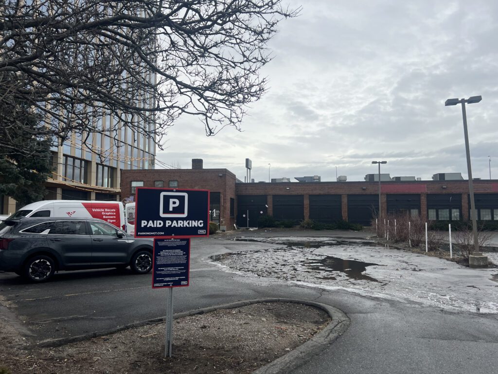 Parking lot with a PMC “Paid Parking” sign and a few parked cars. In the background are brick buildings and trees under a cloudy sky.