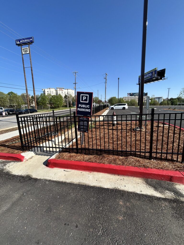 A black metal fence surrounds a landscaped area next to a parking lot. A "Public Parking" sign is attached to the fence. The sky is clear and blue, with some buildings and signs visible in the background.